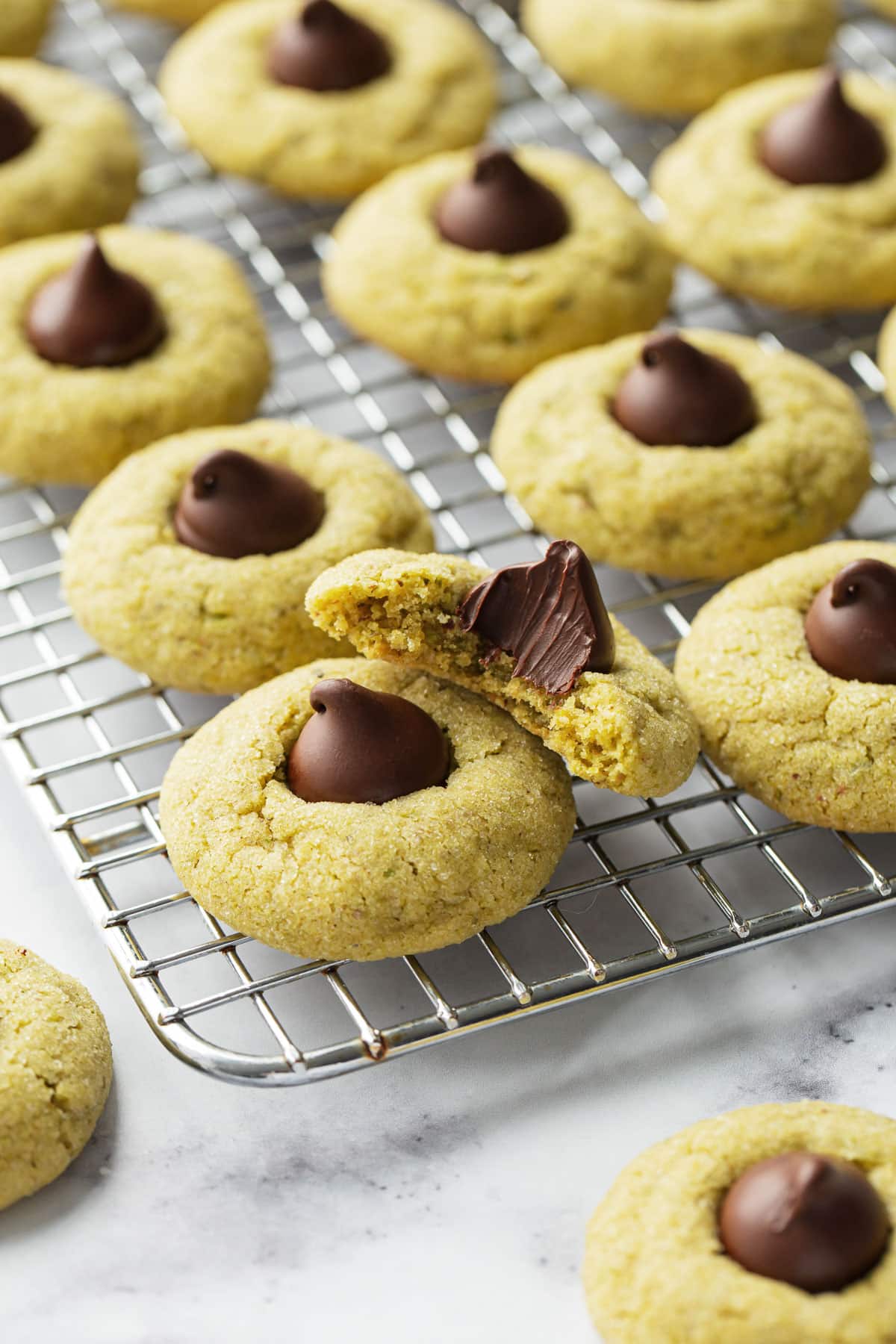 Wire baking rack with rows of Pistachio Butter Blossom Cookies, one bitten cookie resting on top to show the inner texture.