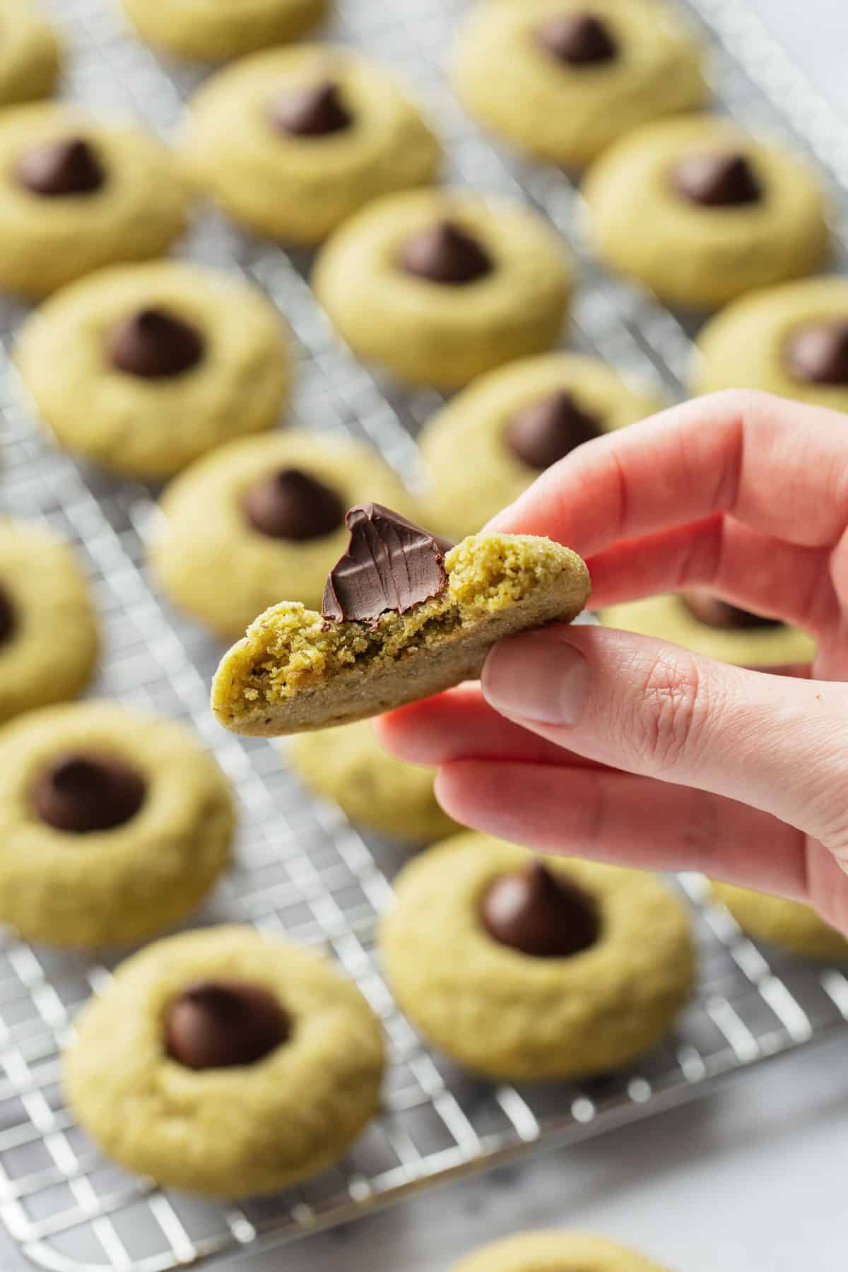 Hand holding a partially bitten Pistachio Butter Blossom Cookie, with a rack with rows of more cookies out of focus in the background.