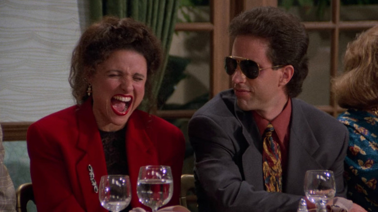 Julia Louis-Dreyfus as Elaine bursts into laughter as Jerry Seinfeld looks on from behind a pair of sunglasses in Seinfeld