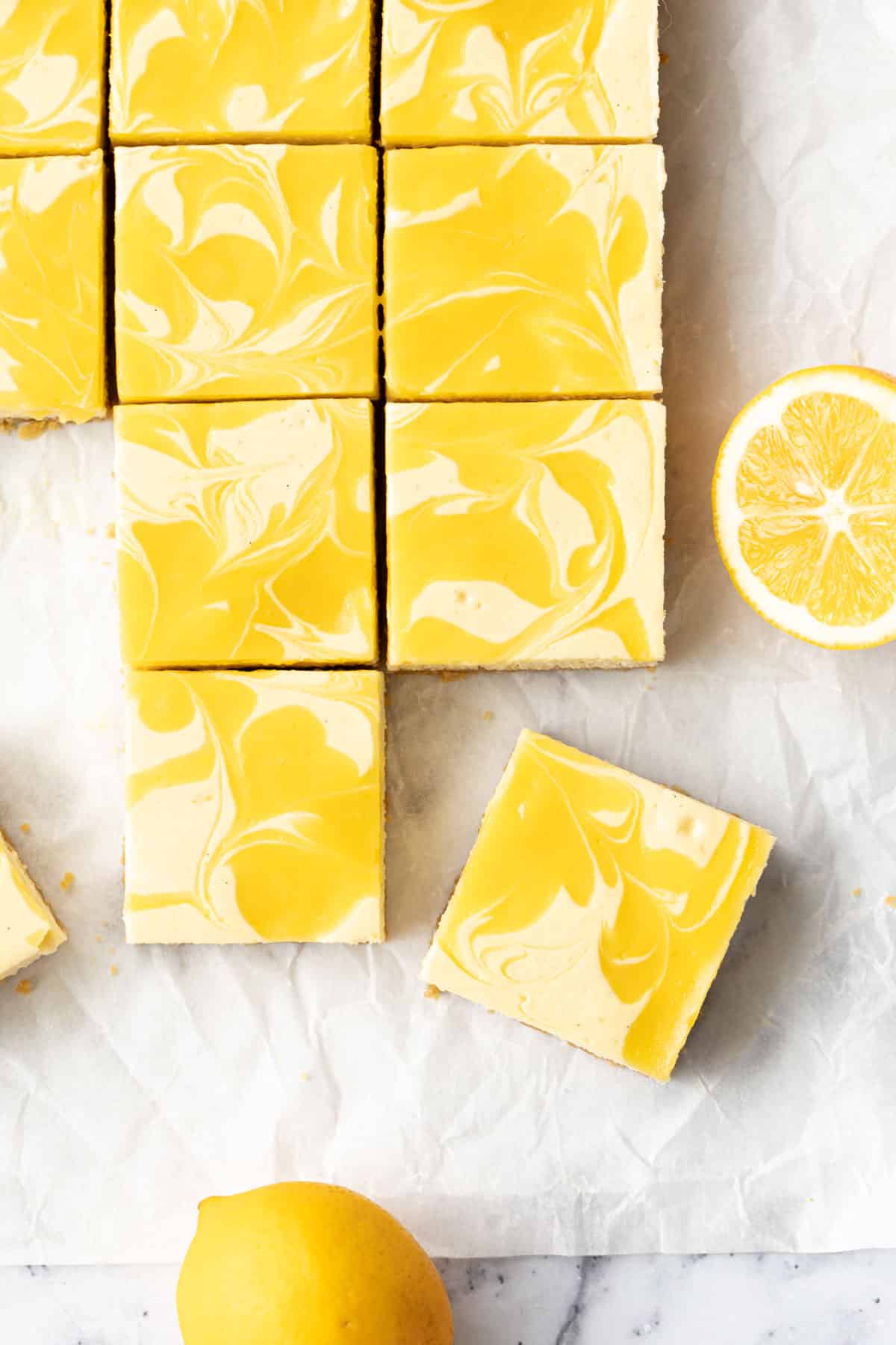 Overhead, squares of Meyer Lemon Cheesecake Bars with swirls of bright yellow lemon curd, on a piece of crinkled parchment with a few whole and cut lemons.