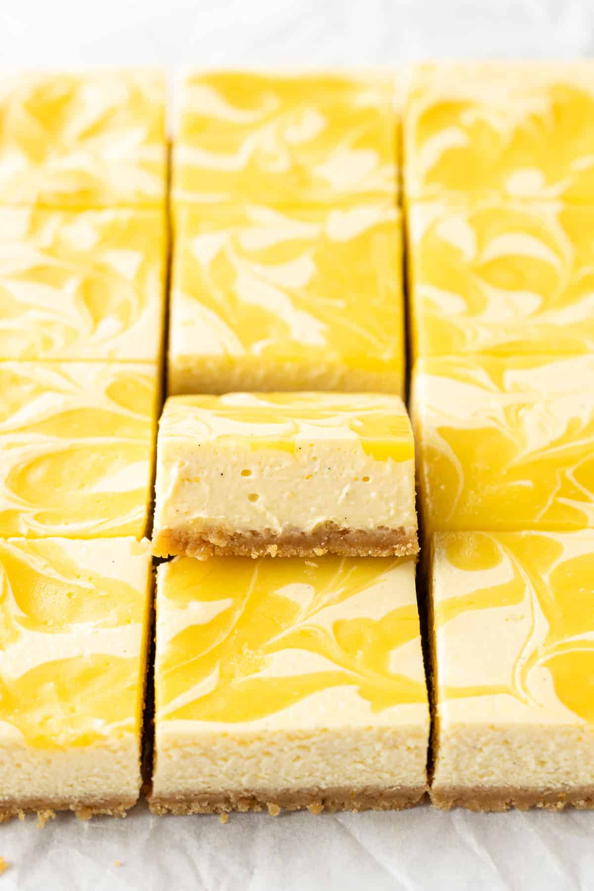 Closeup, squares of Meyer Lemon Cheesecake Bars with swirls of Lemon Curd, one piece tipped upwards to show the layers of crust and cheesecake.