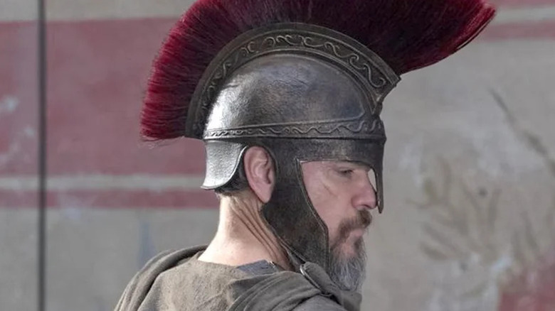 Odysseus wears a metal Roman helmet with red frill and armor while looking pensively over his shoulder in 